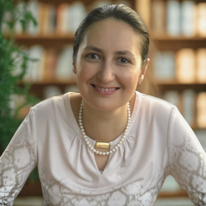 Yana Rulinsky, smiling, wearing a pale top and a pearl necklace, in front of a wall of bookshelves.